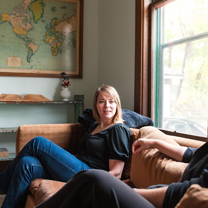 Drei Frauen, vermutlich in ihren Dreißigern bis Fünfzigern, sitzen in einem gemütlichen Wohnzimmer mit einer großen Weltkarte im Hintergrund; sie sind in eine Unterhaltung vertieft, zwei auf einem Sofa und eine in einem Sessel.