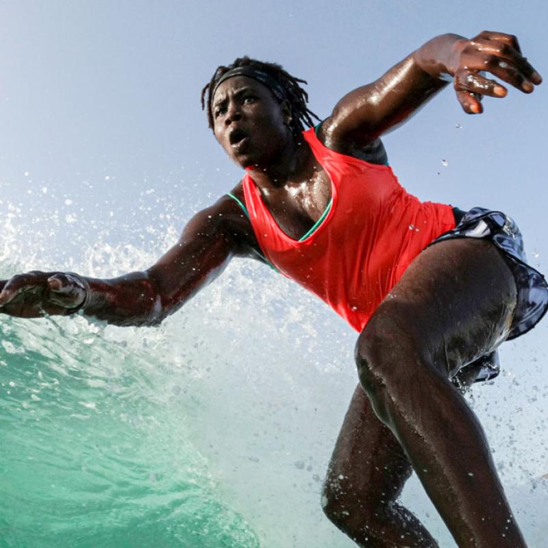 Das Bild zeigt die senegalesische Surferin Khadjou Sambe mit einem  roten Top und einer kurzen Shorts während sie auf einer Welle reitet und ihre Arme zur Balance ausgebreitet hat.