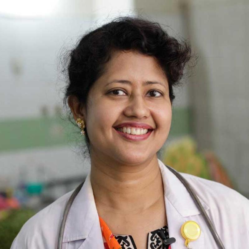 Das Foto zeigt eine bangladesische Ärztin mit einem freundlichen Lächeln. Sie trägt einen weißen Arztkittel, unter dem ein gemustertes Top und ein farbenfroher Schal zu sehen sind, sowie ein Stethoskop um den Hals und ein Namensschild am Kittel. Ihr fröhlicher Ausdruck und die medizinische Ausstattung vermitteln ein Bild von Professionalität im Kontext der fortschreitenden Digitalisierung im Gesundheitswesen Bangladeschs.