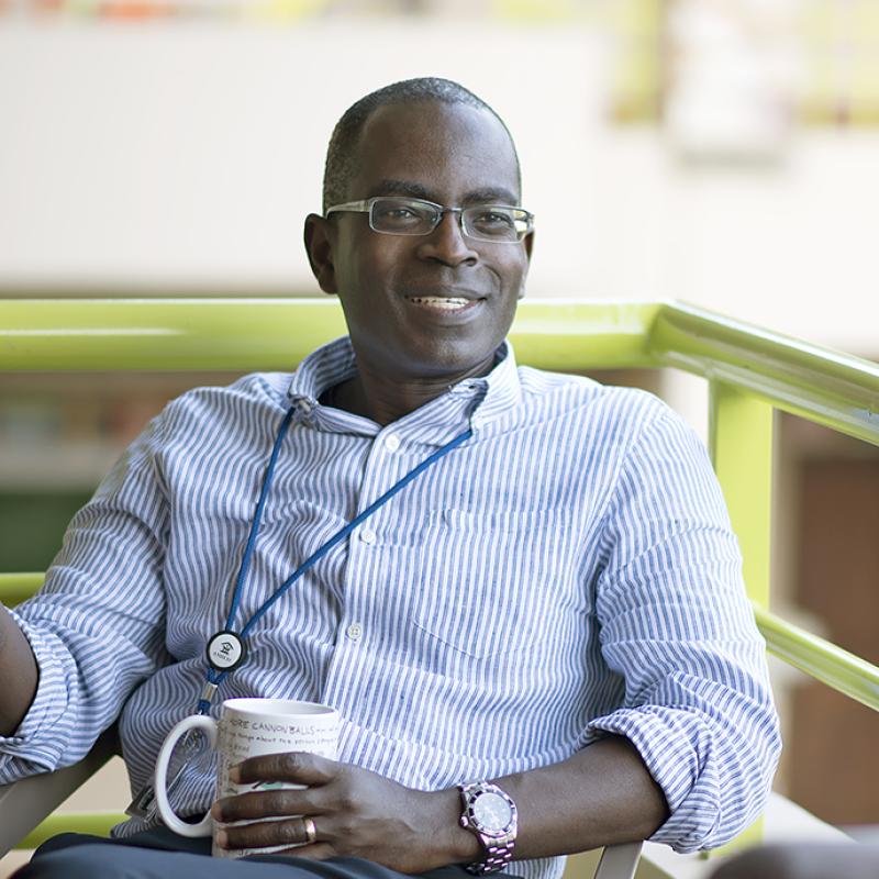 Ein lächelnder Mann mit Brille, gestreiftem Hemd und Ausweis um den Hals sitzt mit einer Kaffeetasse in der Hand auf einer Terrasse, neben ihm ist unscharf der Rücken eines Zuhörers zu sehen.