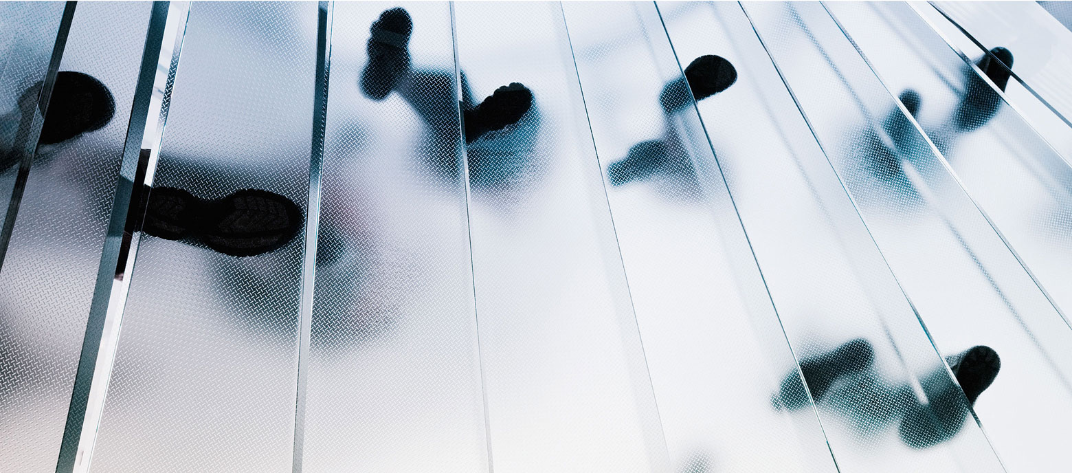 Das Foto zeigt die Schatten mehrerer Personen, die über eine durchscheinende Glas- oder Kunststofffläche laufe. Die Oberfläche ist in Streifen unterteilt, was die Schatten zusätzlich streckt und ein dynamisches Muster erzeugt. Die Perspektive ist von unten nach oben gerichtet, wobei die Füße und Beine der Personen deutlich erkennbar sind.