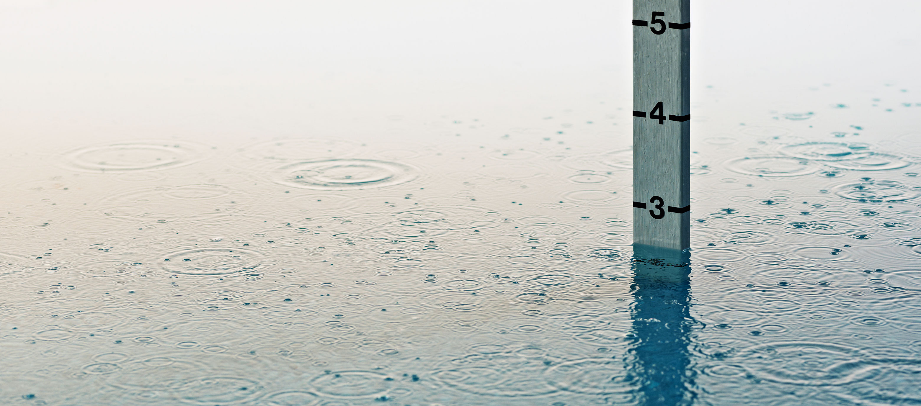 Ein Messtab steht im Wasser, das mit Regentropfen übersät ist und leichte Wellen bildet. Die Wasserhöhe liegt etwas unter der Ziffer 3.