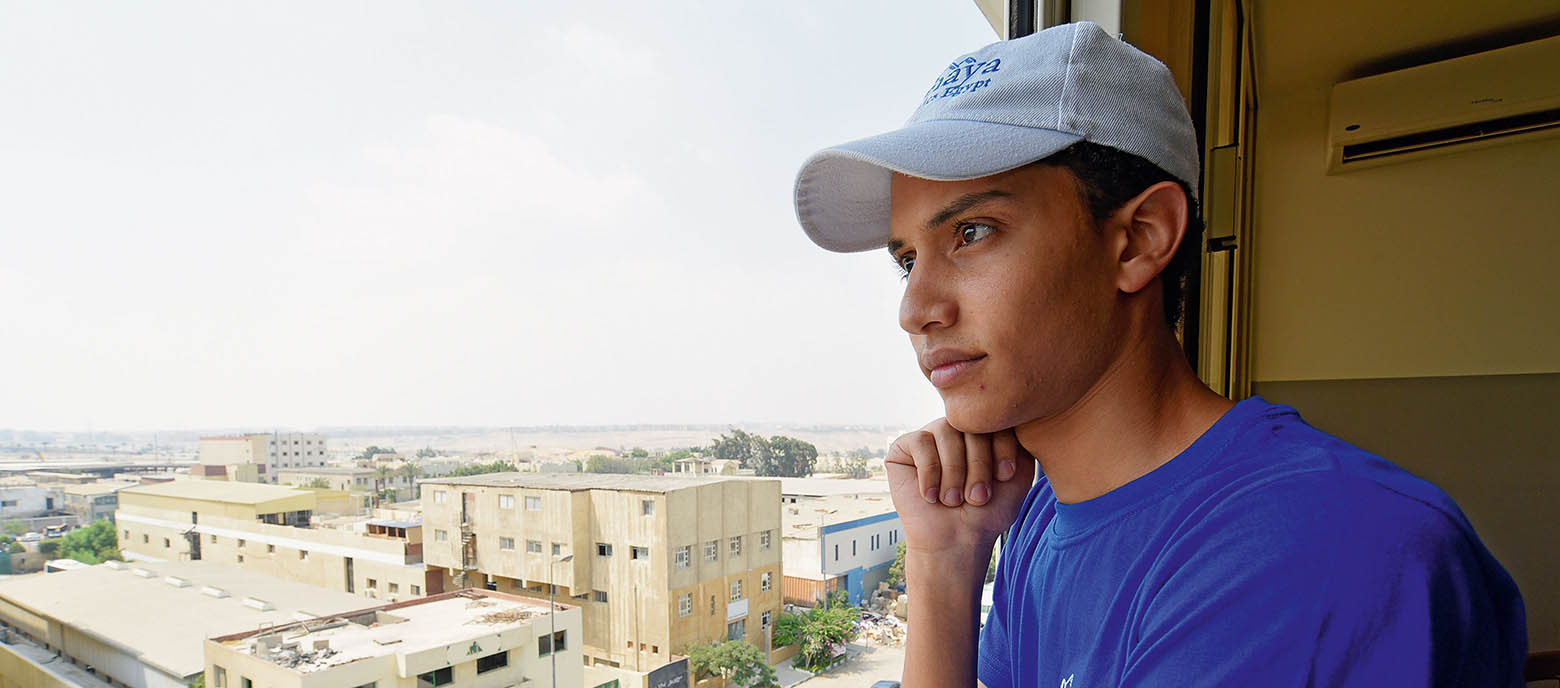 Ein Teenager-Junge mit blauem T-Shirt und weißer Cappy steht nachdenklich vor einem Fenster und stützt sich leicht auf seinem rechten Arm ab. Im Hintergrund sind Gebäude in einer urbanen Umgebung zu erkennen.