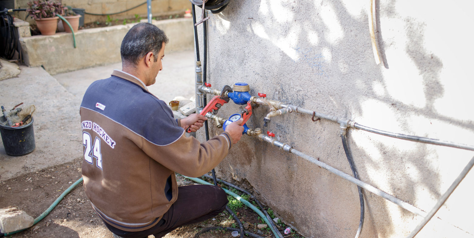 Ein Mann in Palästina ist in der Rückansicht hockend bei der Arbeit an einer Außenwasserinstallation zu sehen. Er benutzt Werkzeuge, um an den Rohrleitungen zu arbeiten, die an einer Außenwand montiert sind.