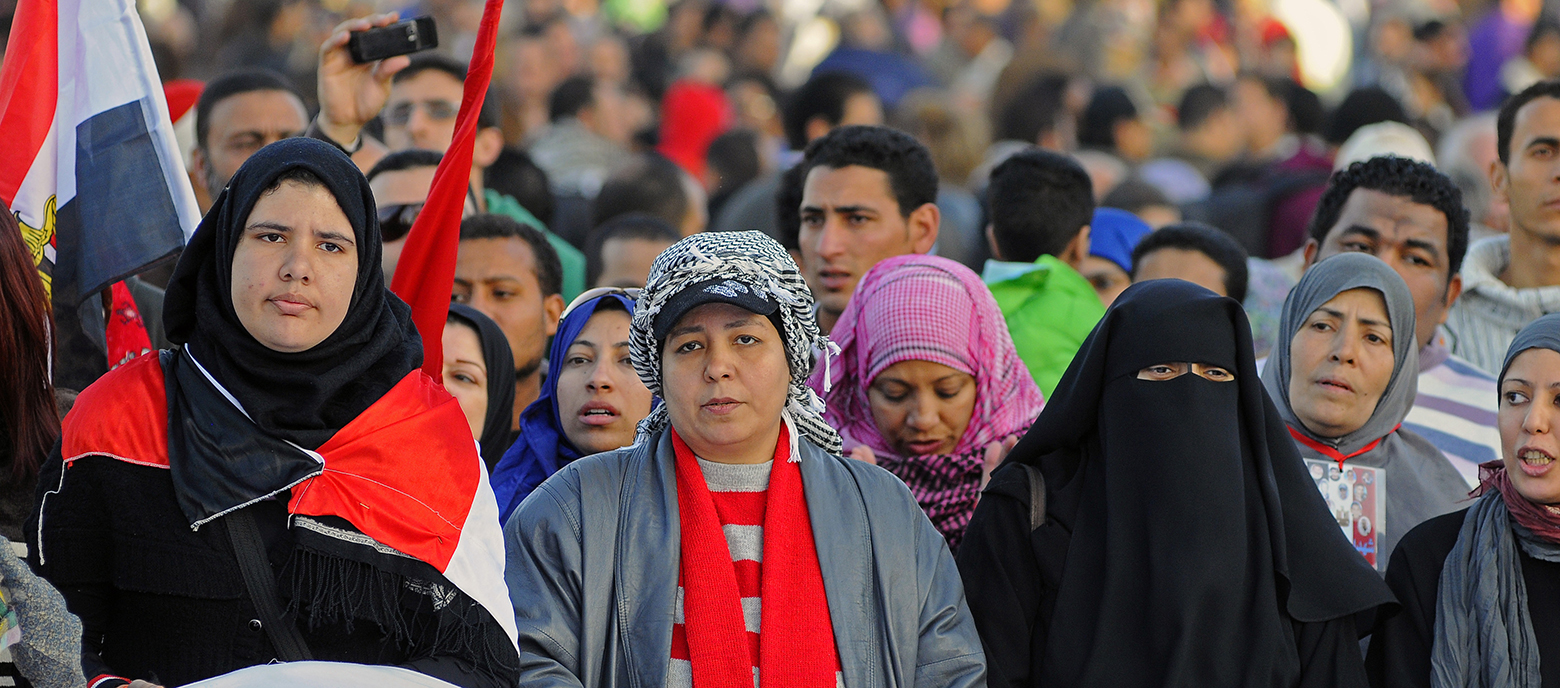 Zwei Frauen, eine mit einem schwarzen Kopftuch und die andere mit einer grauen Strickmütze und einem roten Schal, tragen eine Fahne. Eine dritte Person hebt ein rotes Tuch hoch. Im Hintergrund ist eine Menschenmenge zu sehen.