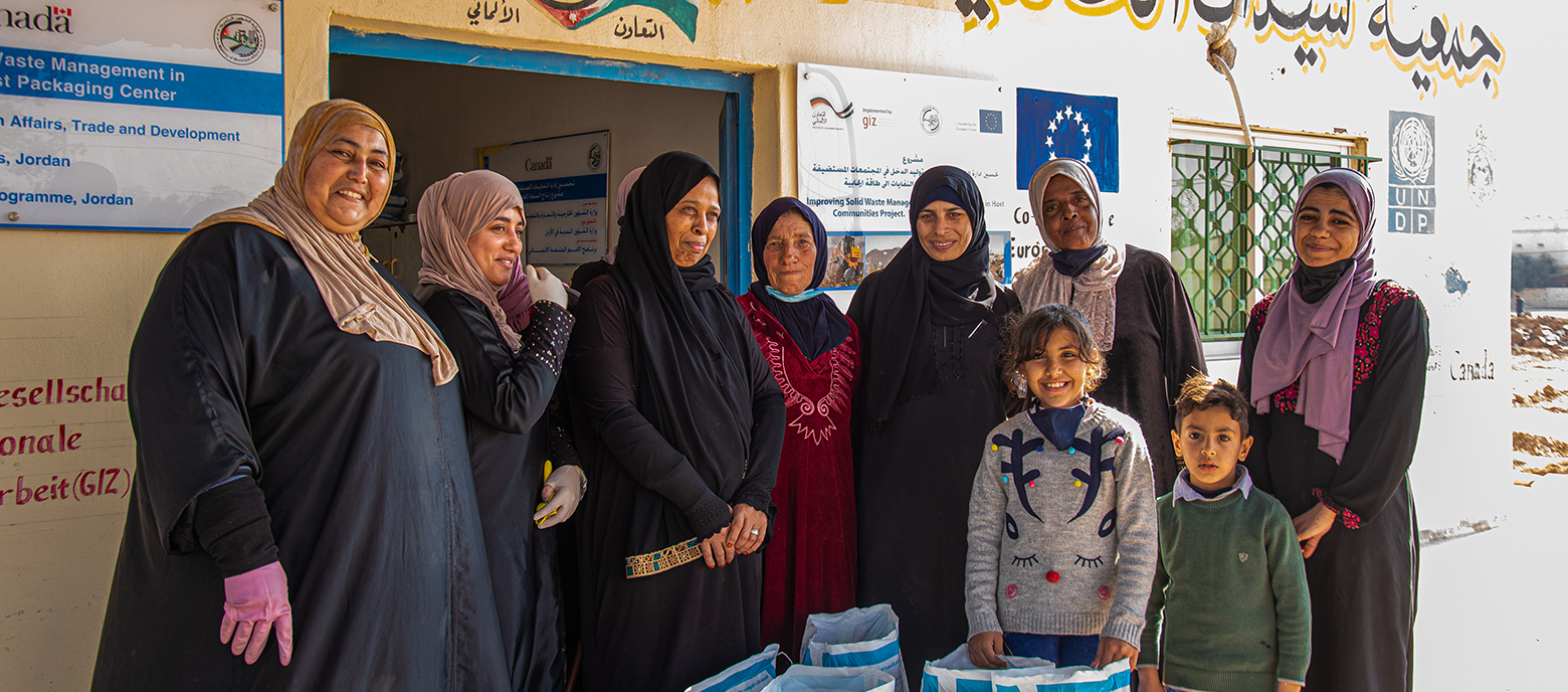 Eine Gruppe lächelnder Frauen und Kinder in traditioneller Bekleidung und Kopftüchern steht vor in einem Entwicklungshilfe-Projektzentrum in Jordanien, umgeben von Kisten für Abfallmanagement.