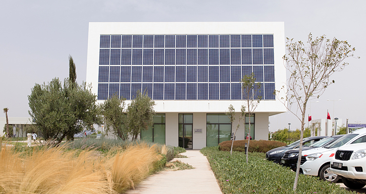 In Frontalsicht ist ein modernes Gebäude zu sehen mit einer Dachfassade voller Solarpaneele. Links vom Haus sind Bäume, Büsche und getrocknete Gräser. Rechts davon sind drei parkende Autos angeschnitten.