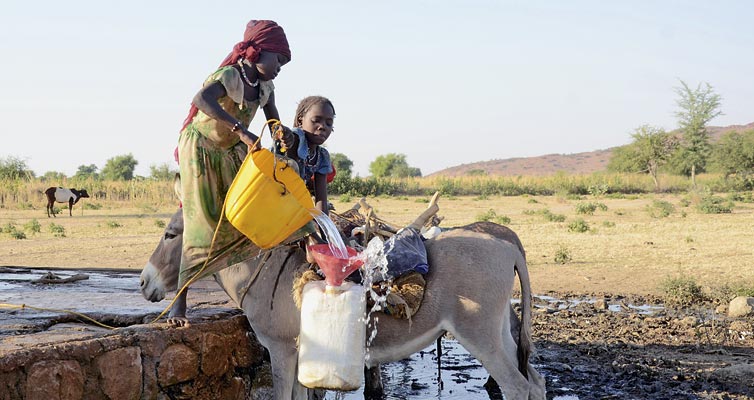 Mutter und Kind in Tschad befüllen Wassergefäße an einem Brunnen in einer ländlichen Umgebug. Das Kind steht auf dem Rücken eines Esels, der die Kanister trägt. Die Frau hilft dabei, den Kanister mit Wasser zu füllen.