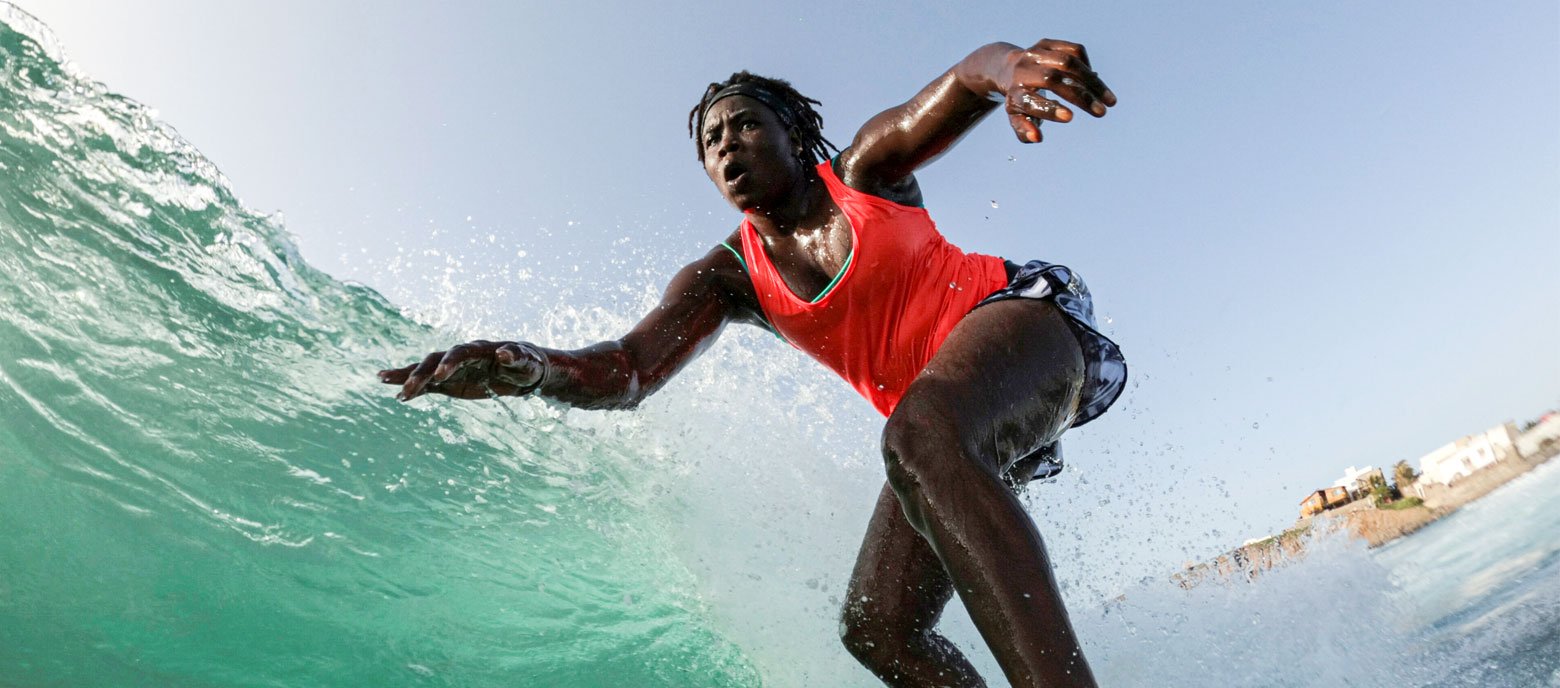 Das Bild zeigt die senegalesische Surferin Khadjou Sambe mit einem  roten Top und einer kurzen Shorts während sie auf einer Welle reitet und ihre Arme zur Balance ausgebreitet hat.