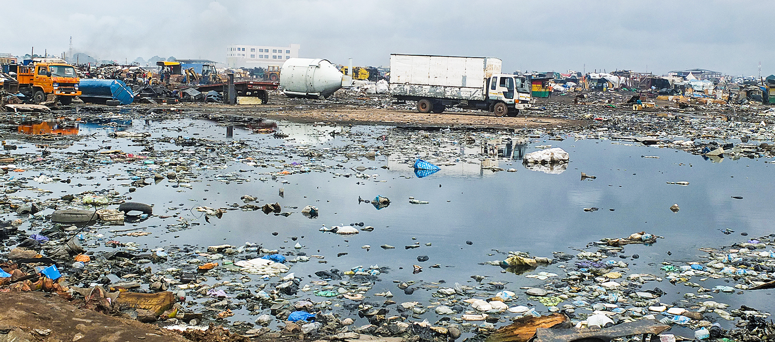 Das Foto zeigt die Elektromüllhalde Agbogbloshie in Accra, Ghana, mit verstreutem Müll, der sich über eine Wasserfläache erstreckt. Im Vordergrund sind Plastikabfälle er erkennen, die das Wasser bedecken und im Hintergrund ist ein weißer LKW neben einem großen weißen Tank von weiteren Abfällen umgeben.