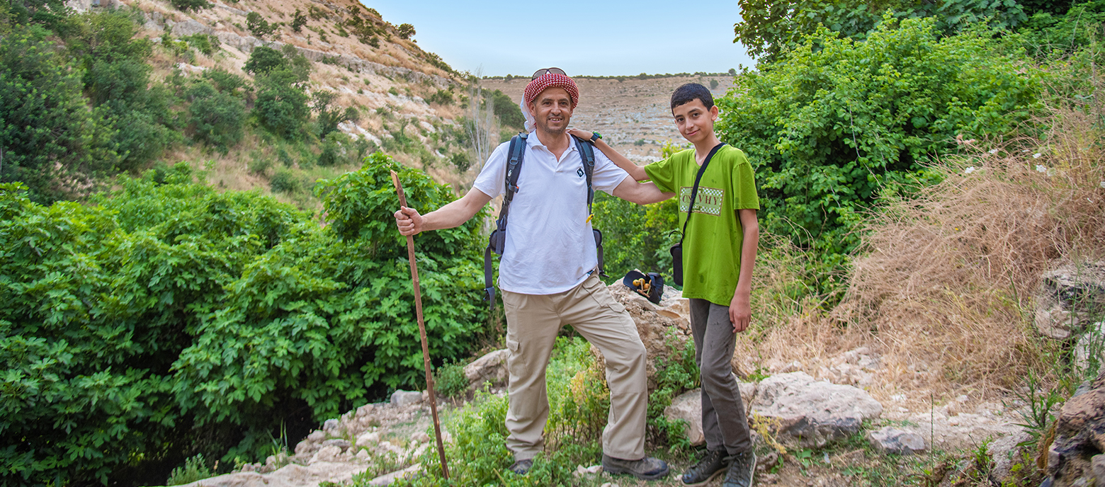 Ein älterer Mann mit einer roten Kopfbedeckung und Wanderstab steht neben einem jungen Jungen in einem grünen T-Shirt. Beide lächeln und der Junge legt seine Hand auf die Schulter des Mannes. Sie befinden sich auf einem Wanderweg, umgeben von Natur und Landschaft.