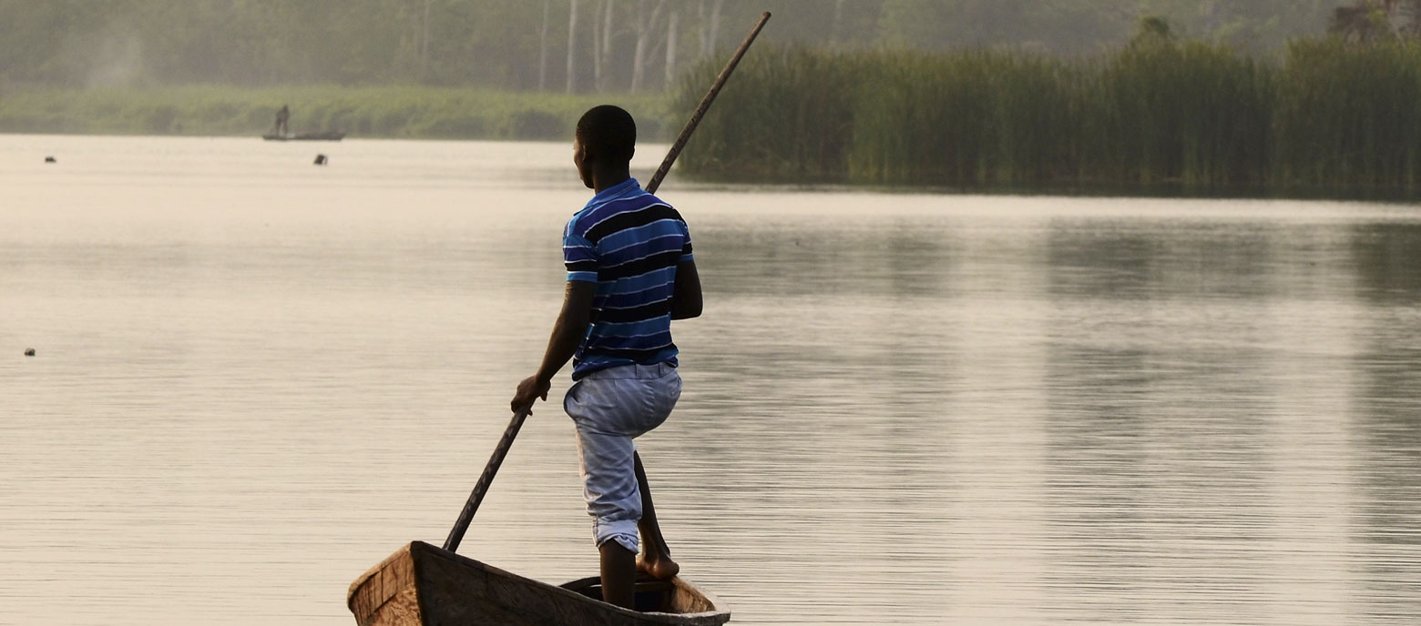 Ein erwachsener Mann steht aufrecht in einem kleinen, traditionellen Holzkanu in Togo und stößt sich mit einem langen Stock vom ruhigen Gewässer ab. Im Hintergrund erkennt man eine Reihe von Bäumen, die am Ufer entlangwachsen.