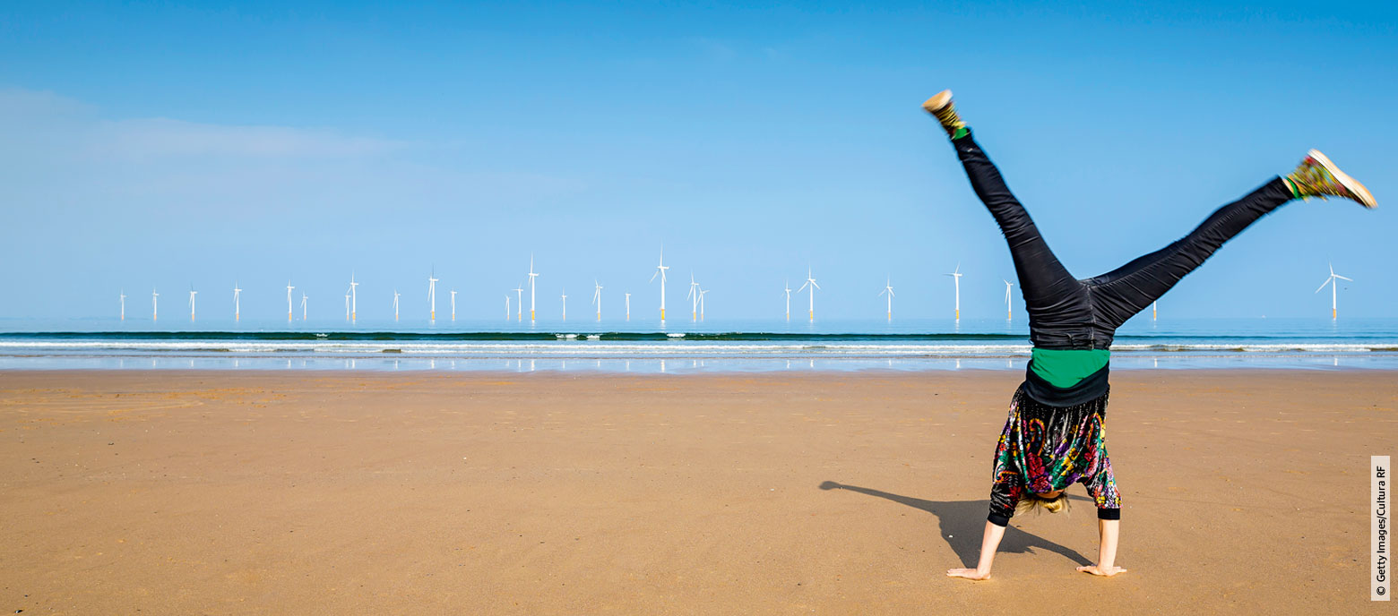 Foto einer Person, die einen Handstand mit gespreizten Beinen auf einem Strand ausführt, im Hintergrund sind mehrere Windturbinen auf dem Meer. Die Person ist von der Kamera weggerichtet, sodass das Gesicht nicht sichtbar ist.