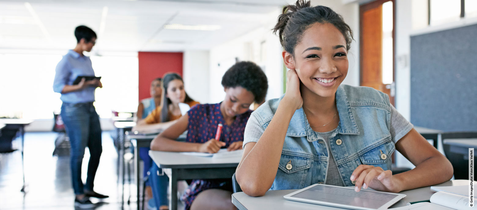 Junge Schülerin mit einem Tablet lächelt in die Kamera, während sie am Tisch in einem Klassenzimmer sitzt. Im verschwommenem Hintergrund arbeiten andere Schüler konzentriert und ein Lehrer geht durch den Raum.