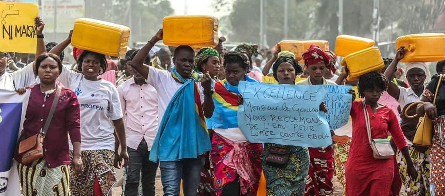 Eine Gruppe von Menschen marschiert auf einer staubigen Straße im Kongo, viele tragen gelbe Wasserkanister auf dem Kopf. Eine Person hält ein Plakat hoch, auf dem auf Französisch steht:  Exzellenz, Herr Gouverneur, wir fordern sauberes Wasser, um gegen Ebola zu kämpfen. Einige tragen traditionelle, bunte Kleidung, andere einfache Alltagskleidung.