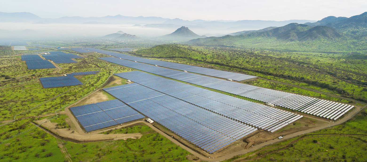 Luftaufnahme eines großen Solarparks mit zahlreichen Photovoltaik-Paneelen, umgeben von grüner Landschaft und Bergen im Hintergrund, die mit leichtem Nebel bedeckt sind.