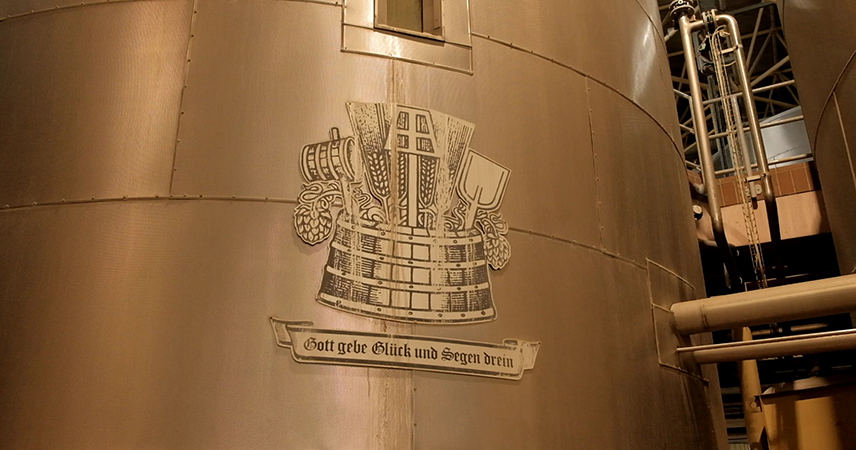 Ein graviertes Emblem auf einer Holzoberfläche, das traditionelle Brauereisymbole darstellt, mit einer Bierkrone in der Mitte, flankiert von Gerstenähren und möglicherweise Brauereiwerkzeugen. Unter dem Emblem befindet sich ein Banner mit der Aufschrift „Gott gebe Glück und Segen drein“.