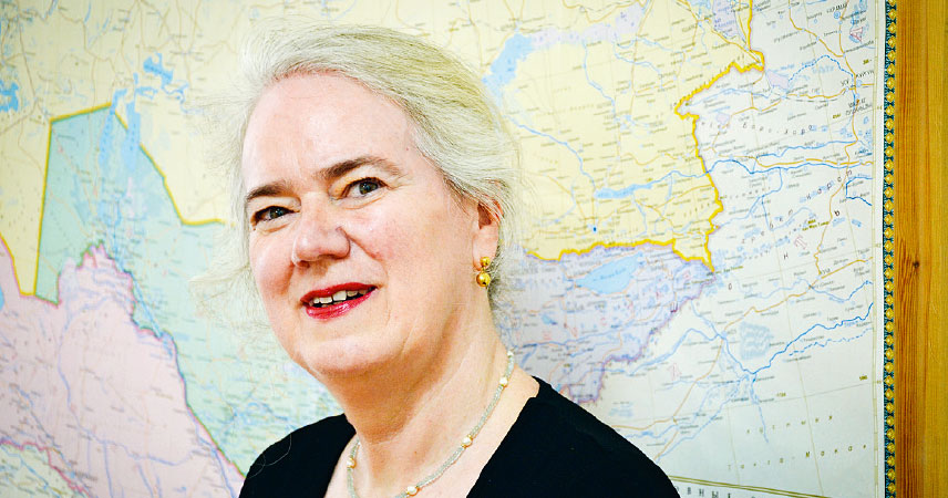 Porträt von Caroline Milow, GIZ Programm-Managerin für Zentralasian. Sie trägt hellblonde bis weiße Haare , eine grüne Kette mit kleinen Perlen und steht vor einer Landkarte, die im Hintergrund zu sehen ist.