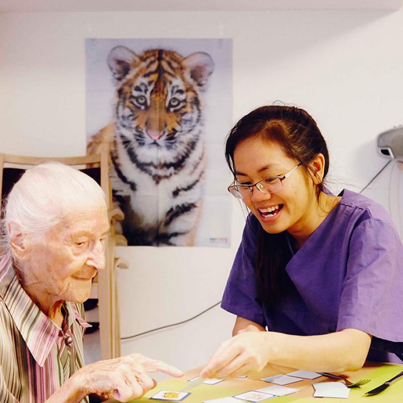 Eine ältere Person und eine jüngere vietnamesische Pflegerin sitzen an einem Tisch und spielen zusammen Karten. Die Pflegerin, erkennbar an ihrer lila Uniform, lacht und scheint der älteren Person beim Kartenspiel zu assistieren. Im Hintergrund ist das Bild eines Tigers an der Wand und Möbelstücke stehen im Raum.