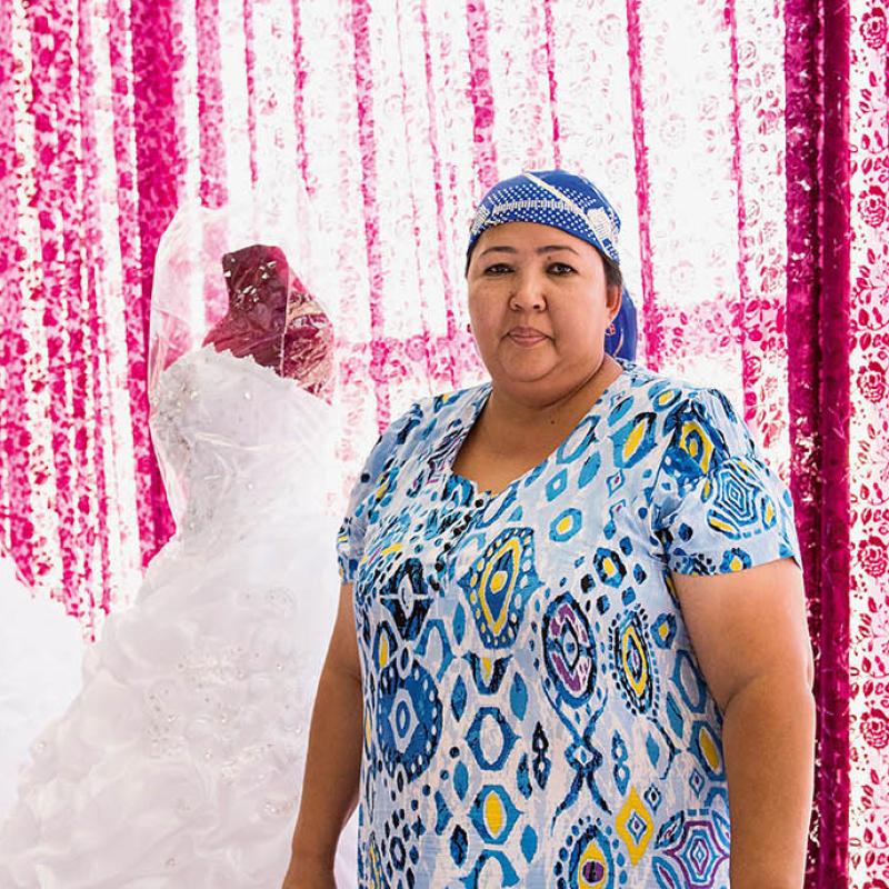 Aufnahme der 40-Jährigen Unternehmerin Shahlo Burhanova in Tajikistan. Sie trägt ein blau-gemustertes Kleid sowie ein passendes Kopftuch dazu. Sie steht aufrecht und schaut ernst in die Kamera. Der Hintergrund sind Hochzeitskleider auf Schaufensterpuppen und ein pink-weißer Vorhang zu sehen.
