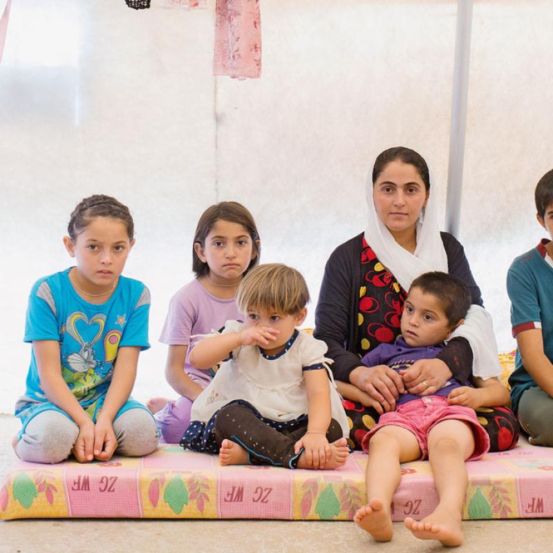 Eine Gruppe von Kindern und einer erwachsenen Frau sitzen auf einer Matratze in einem Flüchtlingslager. Links neben ihnen steht ein einfacher Wasserspender auf einem Hocker.