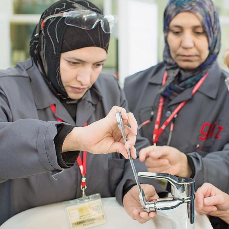 Drei Frauen arbeiten gemeinsam an einem Waschbecken in Jordanien. Die zwei Frauen links tragen Uniformen und Kopftüchern. Während die eine Frau am Waschbecken hantiert, schaut die hintere interessiert zu. Die Frau rechts ohne Kopftuch, in Geschäftskleidung, beobachtet ihre Arbeit. Alle tragen Namensschilder und sind in einem lehrreichen Kontext tätig.