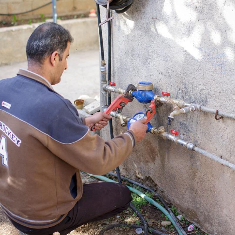 Ein Mann in Palästina ist in der Rückansicht hockend bei der Arbeit an einer Außenwasserinstallation zu sehen. Er benutzt Werkzeuge, um an den Rohrleitungen zu arbeiten, die an einer Außenwand montiert sind.