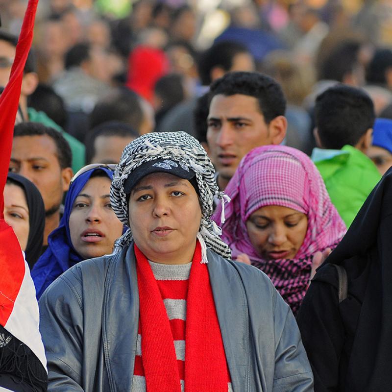 Eine Menschenmenge bei einer Versammlung, im Vordergrund zwei ältere Frauen, die eine Fahne tragen. Die Frau links hält ein Handy hoch, hat ein schwarz-rot-weißes Tuch um die Schultern und trägt ein Kopftuch. Die Frau rechts trägt eine graue Strickmütze und einen roten Schal. Im Hintergrund sind weitere Teilnehmer zu sehen, einige tragen Mützen.