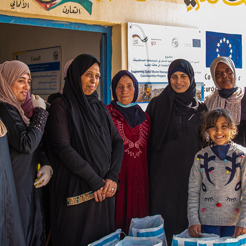 Eine Gruppe lächelnder Frauen und Kinder in traditioneller Bekleidung und Kopftüchern steht vor in einem Entwicklungshilfe-Projektzentrum in Jordanien, umgeben von Kisten für Abfallmanagement.