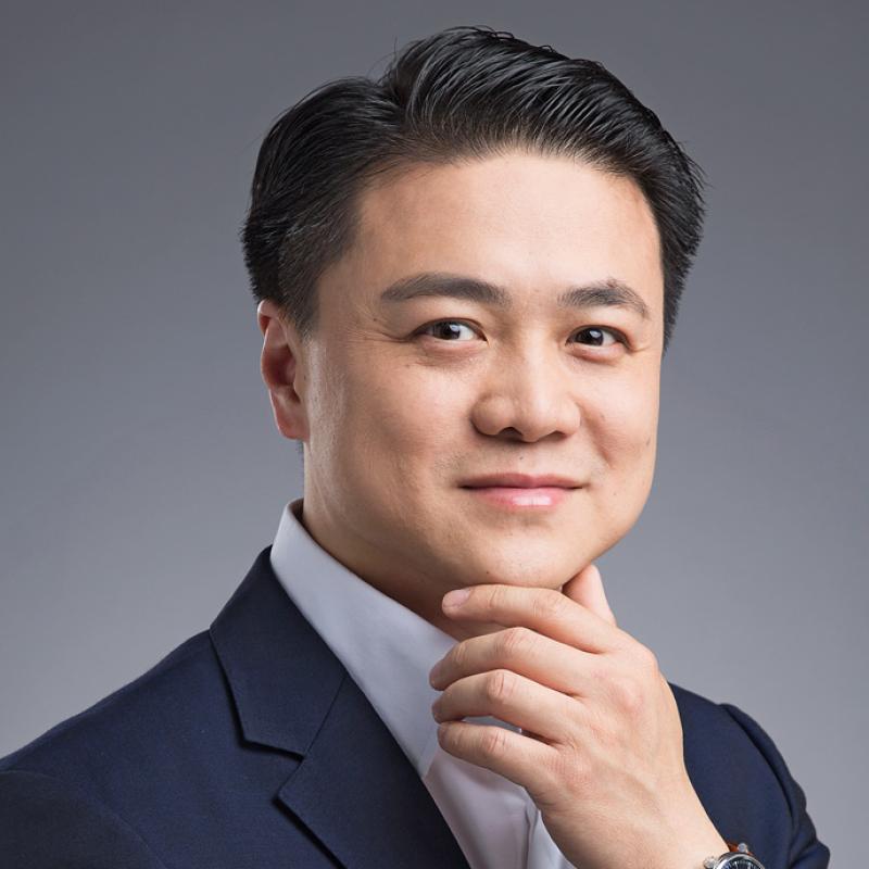 Porträit des jungen Professors Zheng Han in formeller Kleidung. Er lächelt und hält nachdenklich das Kinn mit der rechten Hand. Er trägt einen dunkelblauen Anzug, ein weißes Hemd und eine Uhr am linken Handgelenk. Der Hintergrund ist gleichmäßig grau.
