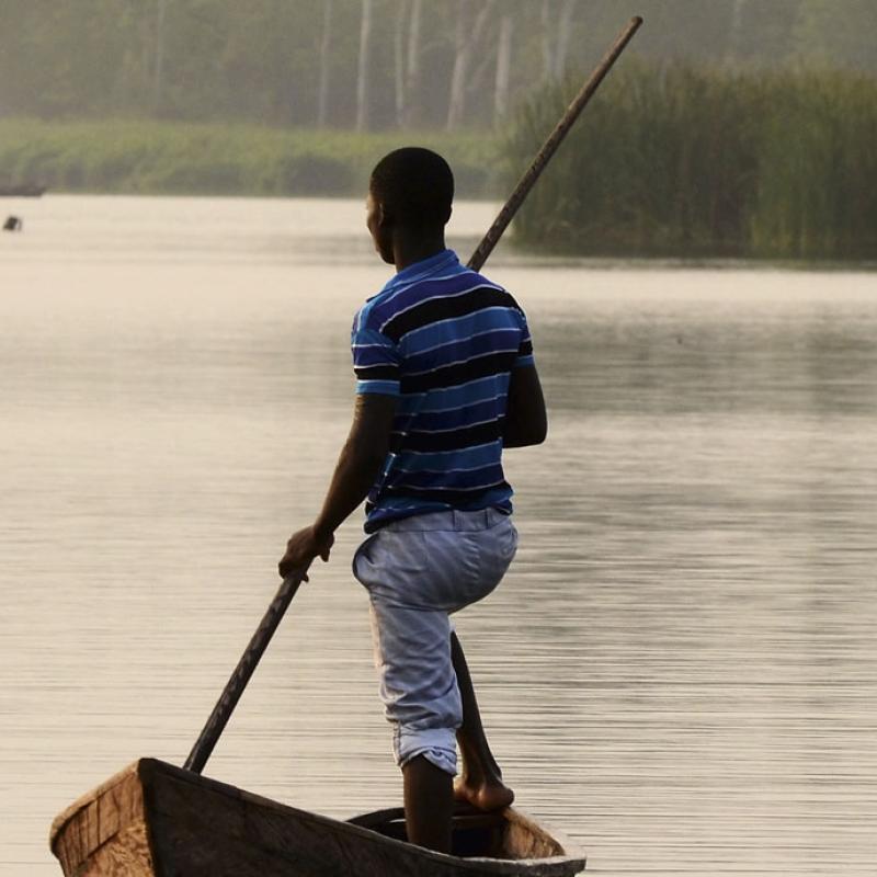 Ein erwachsener Mann steht aufrecht in einem kleinen, traditionellen Holzkanu in Togo und stößt sich mit einem langen Stock vom ruhigen Gewässer ab. Im Hintergrund erkennt man eine Reihe von Bäumen, die am Ufer entlangwachsen.