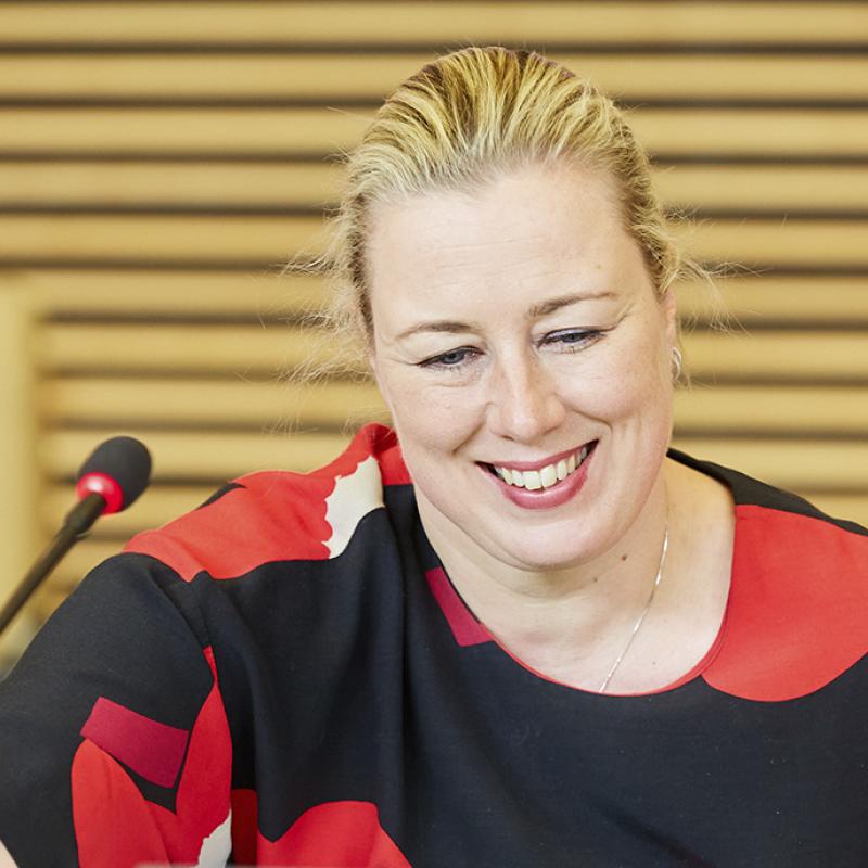 Das Foto zeigt Jutta Urpilainen, die Europäische Kommissarin für internationale Partnerschaften, in einem Konferenzraum mit einem Mikrofon. Sie guckt fröhlich und trägt blonde Haare zum Zopf gebunden und ein Kleid mit auffälligem rot-schwarzen Muster. Der Raum im Hintergrund ist schlicht gehalten mit Holzpaneelen an der Wand.