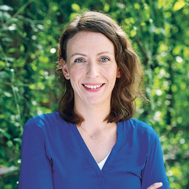 Emilia Huss, eine Klimaexpertin, steht lächelnd im Vordergrund des Bildes. Sie ist mittleren Alters und hat mittellange, lockige braune Haare und trägt eine dunkelblaue Bluse. Der Hintergrund zeigt einen unscharfen grünen Blätterwald.
