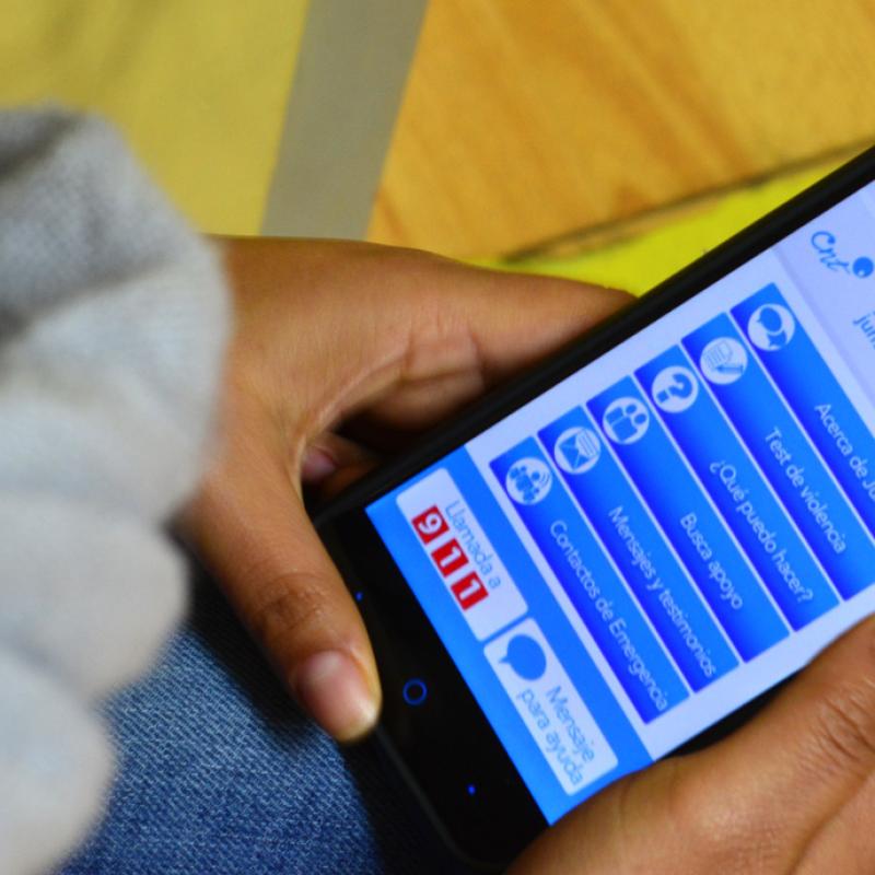 Das Foto zeigt eine Nahaufnahme einer Hand, die ein Smartphone hält, auf dessen Bildschirm eine Anwendung mit mehreren Icons und Text in blauen Farbtönen geöffnet ist. Man kann das obere Ende einer gelben Linie auf dem Boden erkennen, was darauf hindeutet, dass die Person auf dem Boden sitzt oder steht.