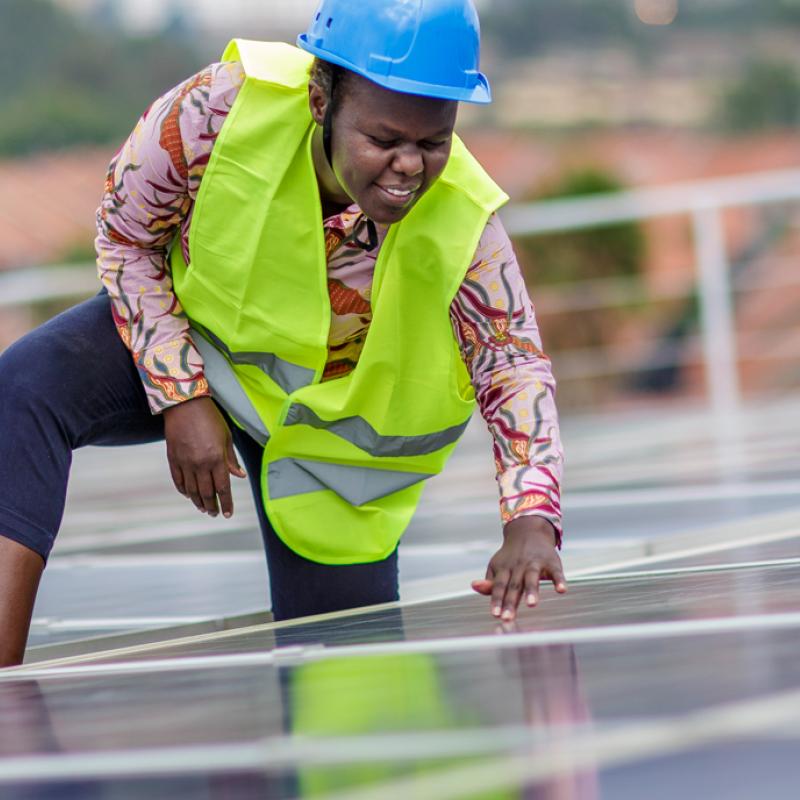 Eine schwarze Frau mittleren Alters mit blauem Helm und neongelber Sicherheitsweste beugt sich über Solarpaneele auf einem Dach und inspiziert diese. Im Hintergrund sind unscharf weitere Dächer zu erkennen. 