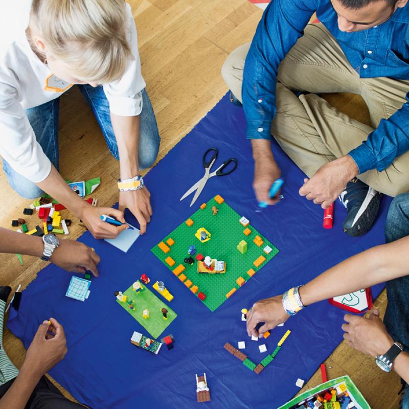 Vier Personen sitzen auf dem Boden und arbeiten gemeinsam an einem Projekt, umgeben von bunten Bausteinen. Sie befinden sich auf einem blauen Tuch, auf dem ein grünes Quadrat mit Bausteinen zu sehen ist. Eine Person schneidet ein Band, während die anderen Bausteine sortieren und zusammenfügen.