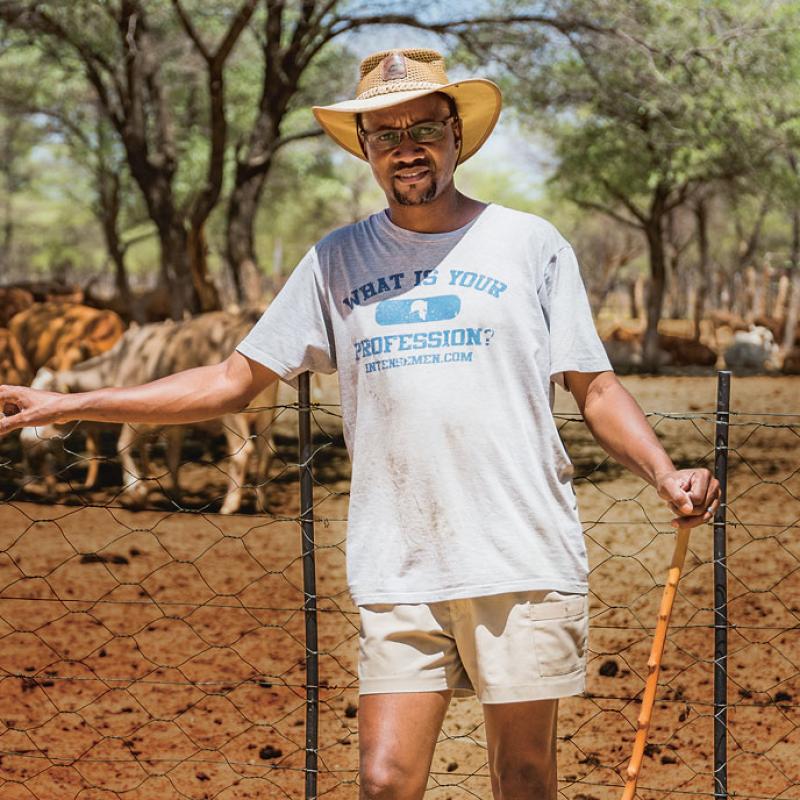 Namibischer Bauer vor einem Zaun stehend. Er trägt ein T-Shirt, eine helle Hose und einen Hut und schaut in die Kamera. Im Hintergrund sind unscharf Kühe und eine ländliche Umgebung zu erkennen.