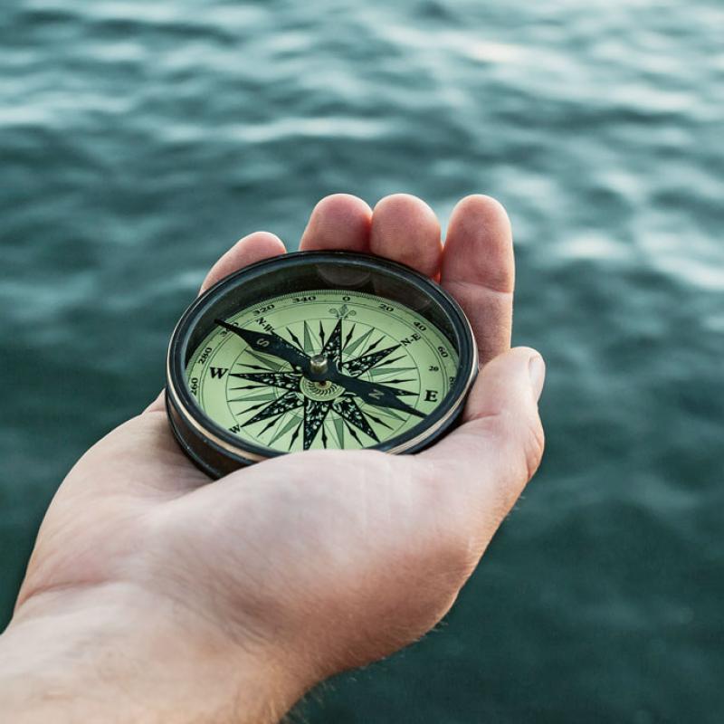 Nahaufnahme einer Hand, die einen Kompass vor verschwommenem Meer im Hintergrund hält.