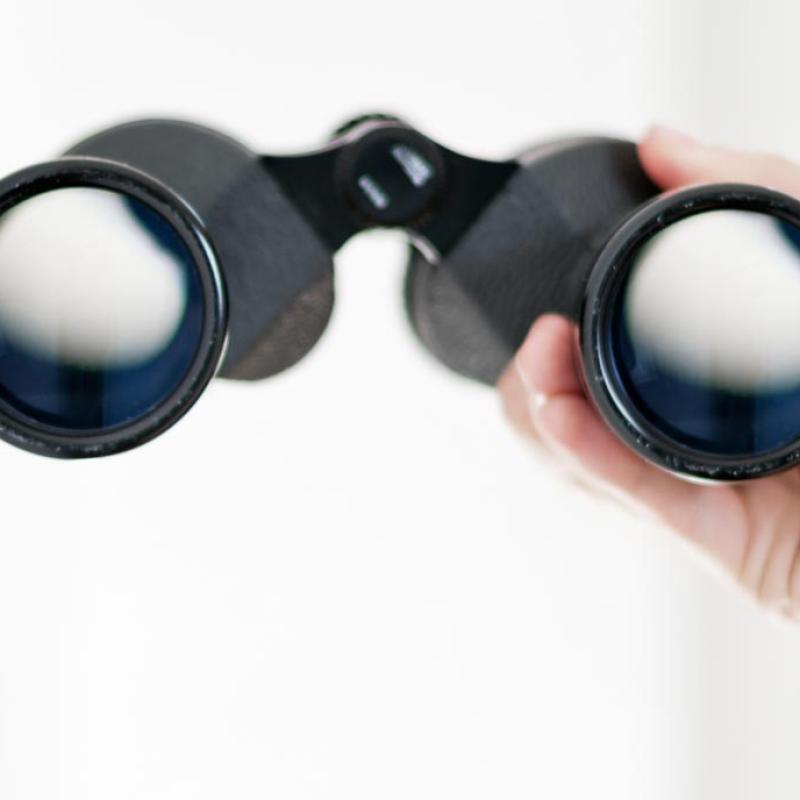 Eine Hand hält ein Fernglas vor einem hellen weißen Hintergrund. Das Fernglas ist schwarz und erscheint im Fokus.