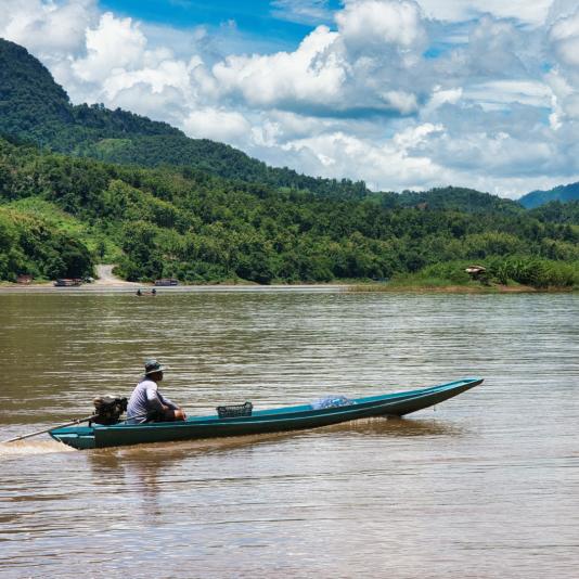 Ein Mann in einem langen, schmalen Boot bewegt sich über einen trüben Fluss, umgeben von grünen Hügeln und einem wolkenverhangenen Himmel. Der Mann, der am hinteren Ende des Bootes sitzt, trägt einen Hut und ein ein helles Oberteil. 