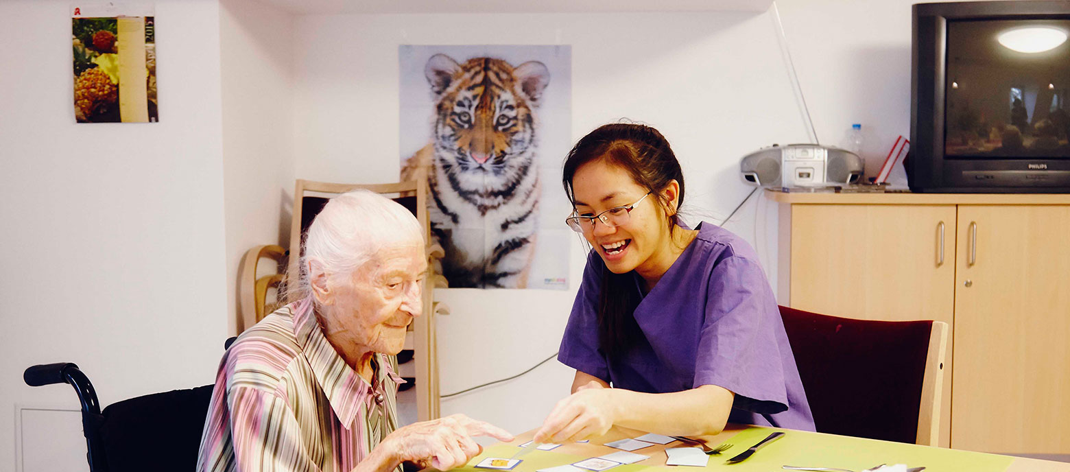 Eine ältere Person und eine jüngere vietnamesische Pflegerin sitzen an einem Tisch und spielen zusammen Karten. Die Pflegerin, erkennbar an ihrer lila Uniform, lacht und scheint der älteren Person beim Kartenspiel zu assistieren. Im Hintergrund ist das Bild eines Tigers an der Wand und Möbelstücke stehen im Raum.