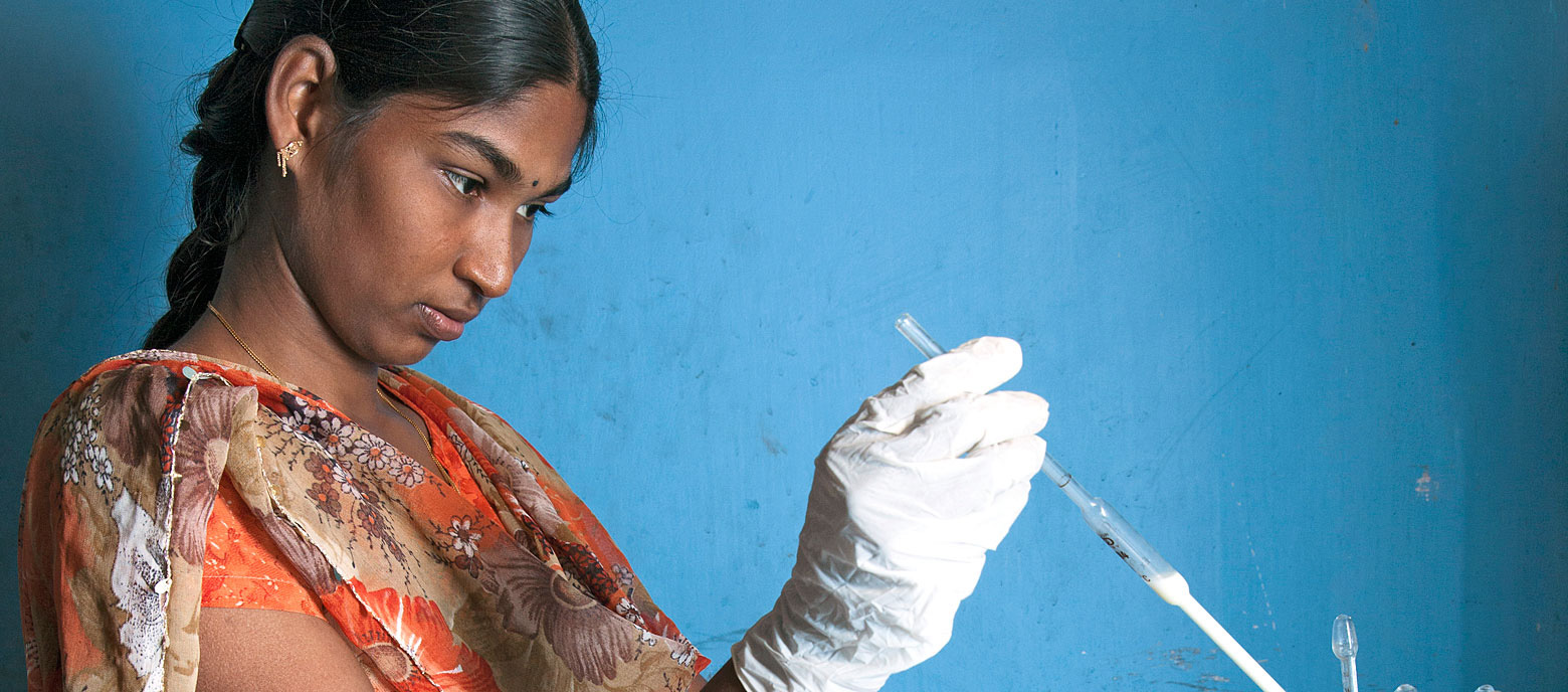 Eine Frau in traditioneller indischer Kleidung konzentriert sich auf das Befüllen einer Spritze. Sie trägt medizinische Handschuhe und steht links im Bild vor einer blauen Wand.