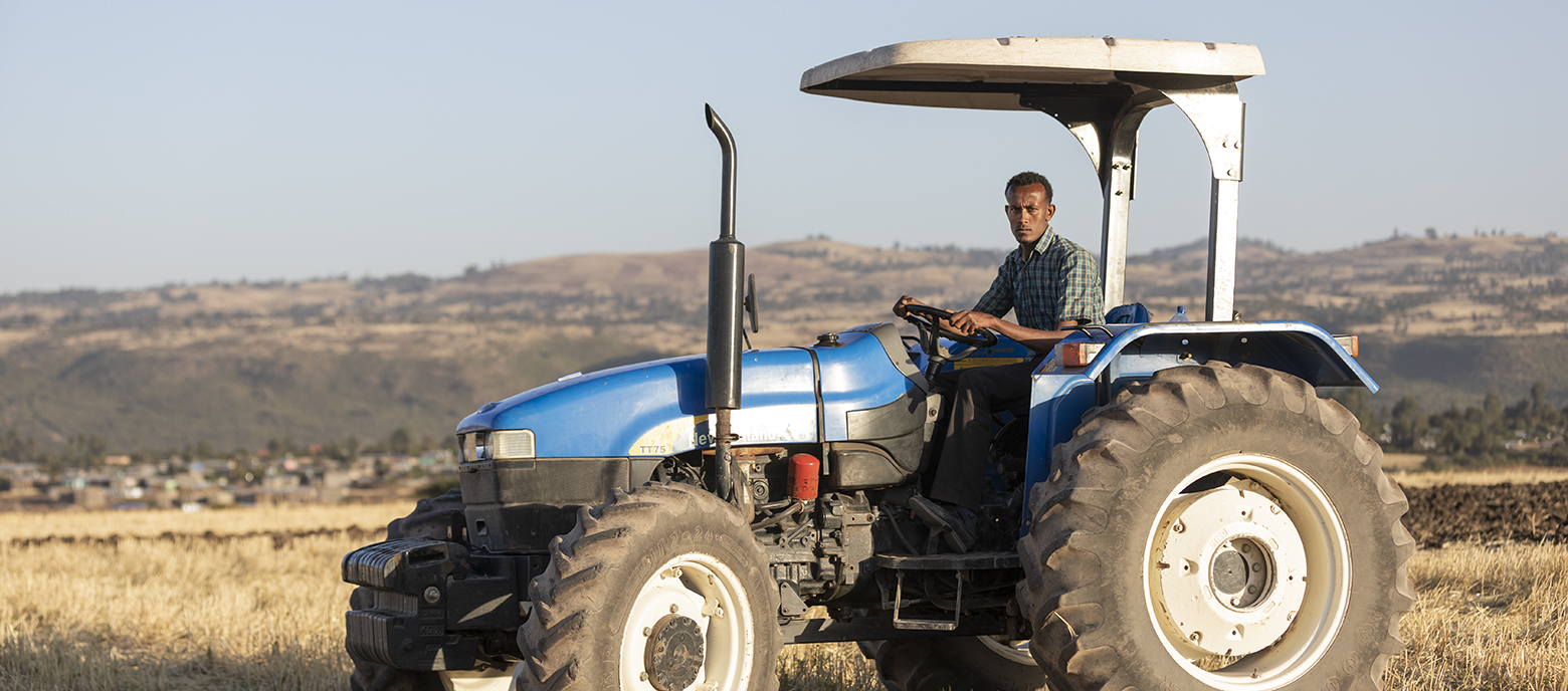 Ein Bauer in Äthiopien fährt einen blauen Traktor über ein trockenes Feld mit Hügeln und einer kleinen Siedlung im Hintergrund. Der Himmel ist klar, und der Bauer schaut konzentriert in die Kamera.