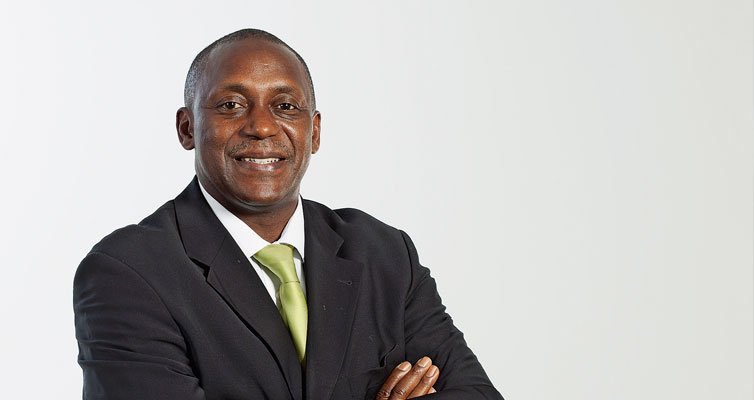 Portrait von Kandeh Yumkella, der in einem dunklen Anzug mit einer hellgrünen Krawatte gekleidet ist. Er steht mit verschränkten Armen und lächelt direkt in die Kamera. Sein Auftreten ist professionell und selbstsicher.