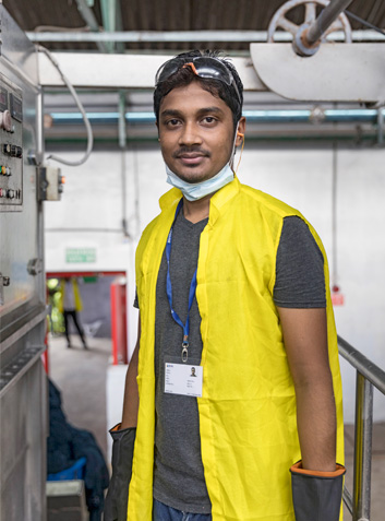 Ein Mann steht in einer Textilfabrik in Bangladesch und blickt in die Kamera. Er trägt eine gelbe Sicherheitsweste, eine heruntergezogene Gesichtsmaske um den Hals und eine Schutzbrille auf dem Kopf sowie ein dunkles T-Shirt und einen Ausweis um den Hals. Im Hintergrund sind Maschinen und ein weiterer Mensch in Sicherheitskleidung zu erkennen.