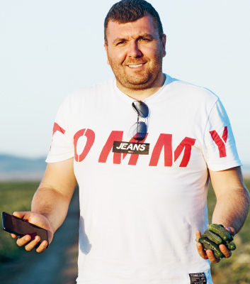 Ein Mann steht draußen bei Tageslicht und hält ein Smartphone in der Hand. Er trägt ein weißes T-Shirt mit der Aufschrift 