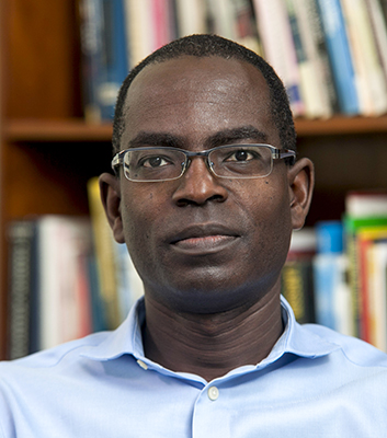 Patrick Awuah mit Brille und blauem Hemd blickt direkt in die Kamera. Im Hintergrund sind Bücherregale sichtbar.