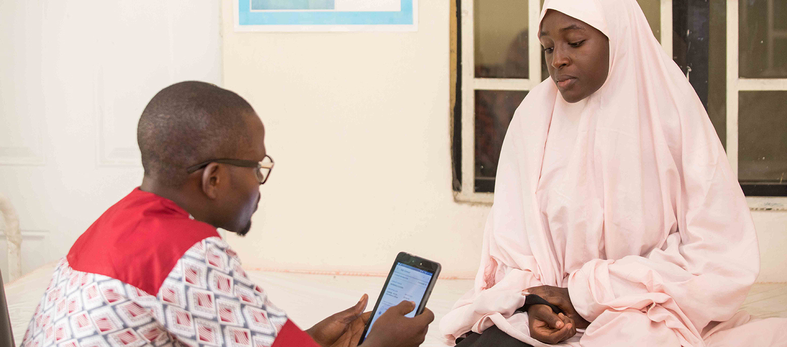 Ein Mann mit Brille in traditioneller nigerianischer Kleidung  zeigt einer Frau in einem hellrosa Hijab etwas auf einem Smartphone. Sie sitzen einander in einem hellen Raum gegenüber, die Frau hört aufmerksam zu.