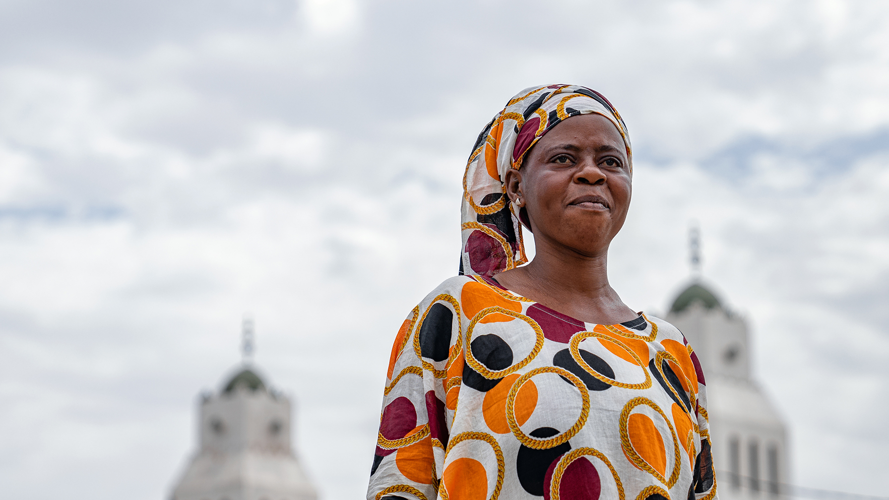 Eine Frau in einem bunten traditionellen Kleid steht vor zwei weißen Türmen mit grünen Kuppeln unter einem bewölkten Himmel.