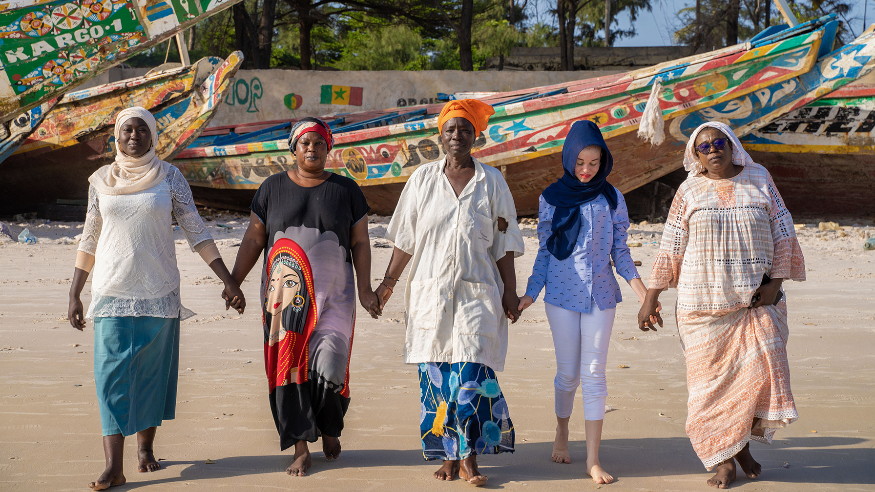 5 women walk hand in hand on the beach with colorful boats in the background